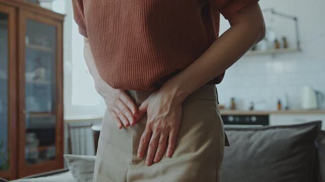 Unrecognizable woman standing at home, bending and holding her stomach as feeling painful cramps. Close-up view, midsection shot