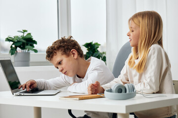 Fototapeta premium Happy children studying together online at home, using laptops and sitting at a table in the living room The boy and girl, both wearing headphones, are concentrated and engaged in their elearning