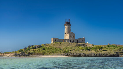 An old dilapidated lighthouse on an island in the ocean. A tall tower against a clear blue sky. The green grass on the hill. Turquoise sea water in the foreground. Mauritius. Île au Phare 