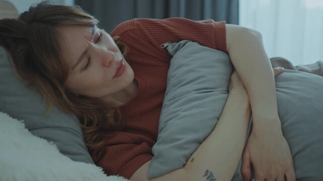 Young woman lying down on bed at home, hugging pillow and wincing as suffering from cramps in stomach
