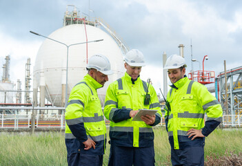 Three engineer or technician workers stand in front of petrochemical tank in factory and they discuss with tablet together.