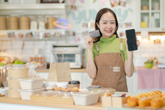 Young chinese woman employee holding data phone and credit card at cafe coffee.Smiling asian barista girl, wears apron, shows credit card machine for processing payment standing in coffee shop.