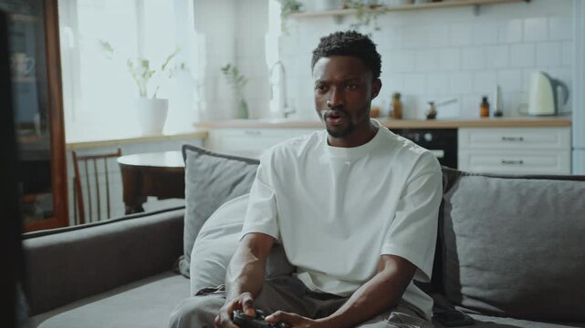 Young interested Black man sitting on sofa and using controller as playing video game on console during the day at home