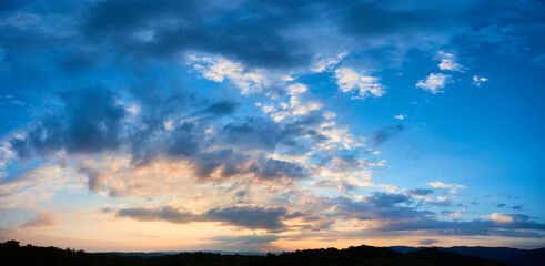 Fototapeta premium Breathtaking sky filled with dramatic clouds transitions from deep blue to soft pastels as sun sets. Horizon lined with silhouetted hills and forests, adding depth to serene and expansive landscape.
