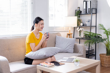 Happy young asian woman drinking coffee relaxing on sofa at home. Smiling female enjoying resting sitting on couch in modern living room.
