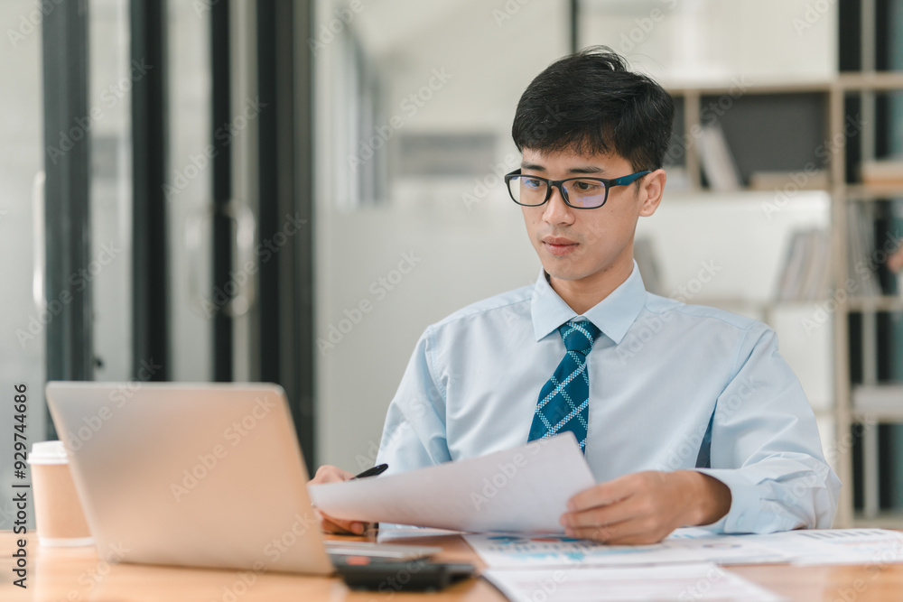 Wall mural a man wearing a blue shirt and a tie is sitting at a desk with a laptop open in front of him. he is 