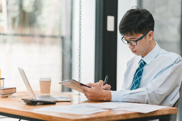 A man in a blue shirt and a blue tie is sitting at a desk with a laptop and a clipboard. He is writing on the clipboard with a pen. The scene suggests a professional setting