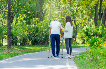 back view of daughter help disability father walking in the park,a young adult woman supporting a senior man using a walker to walk in the park