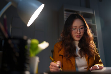 Young professional is working late, illuminated by a desk lamp and concentrating on taking notes