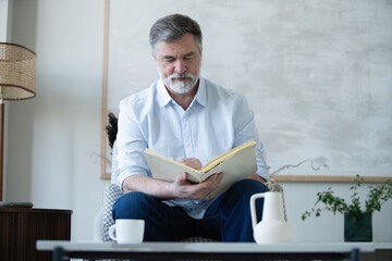 Senior Man Reading Book Sitting On Couch At Home. Retired Male Enjoying Reading New Novel Or Business Literature On Weekend