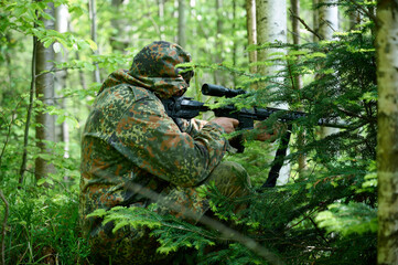 Hunter with rifle wearing camouflage suit sitting in ambush. Camouflaged sniper in the forest in ambush. Military man hide in the woods with rifle.