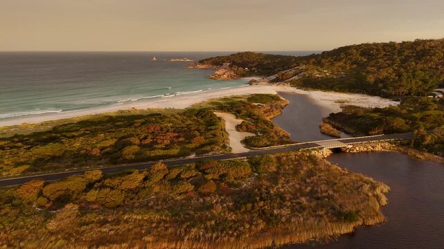 Aerial flyover lake, island with street and sandy beach during sunset time. Clear ocean and beautiful coastline at Bay of Fires. Wide shot. Tasmania, Australia.