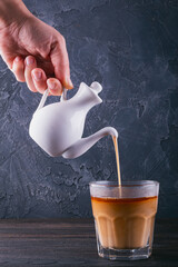 Pouring a rich and creamy coffee beverage from a pristine white teapot into a clear glass cup