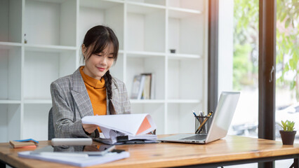 A positive Asian businesswoman is reviewing the financial report while working at her desk.