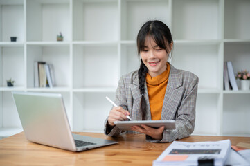 A charming, positive Asian businesswoman is using a digital tablet at her desk in the office.