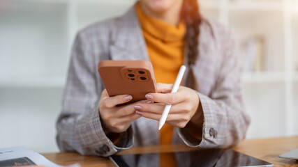 An Asian businesswoman is holding a stylus pen and using a smartphone at her desk in the office.