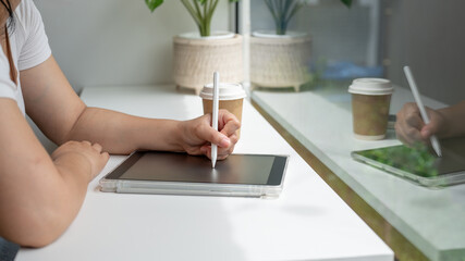 A woman holding a stylus pen, writing on a digital tablet, working remotely from a coffee shop.