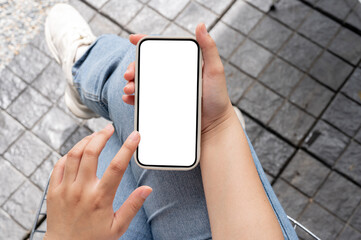 Top view image of a woman in jeans sitting outdoors, using her smartphone, touching on the screen.