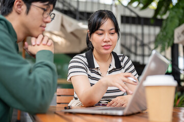 A positive Asian woman is working on a co-project with her friend, showing something on her laptop.