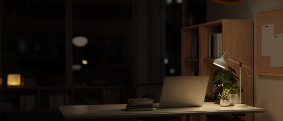 A modern dark office at night, a laptop and accessories on a desk, illuminated by a table lamp.