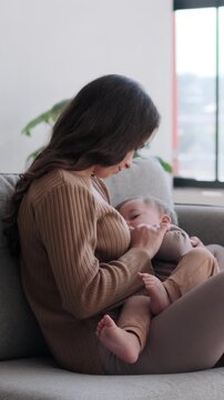Caring Caucasian mother breastfeed toddler son on living room sofa at home. Motherhood, childcare and family love concept. Vertical video.
