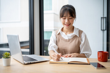 A beautiful, positive Asian woman writing in a book, working at a desk indoors, doing homework.