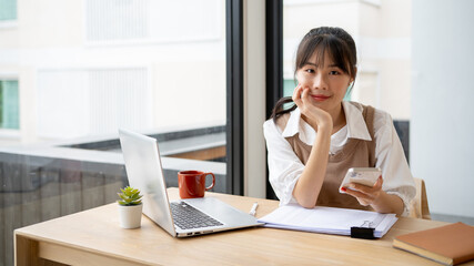 A beautiful Asian woman is sitting at a desk with a smartphone in her hand and smiling at the camera