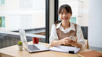 A beautiful Asian woman is sitting at a desk with a smartphone in her hand and smiling at the camera