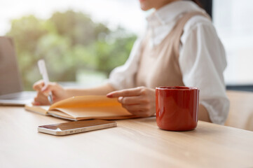 A cropped image of a woman writing in a book, working at her desk in the office.