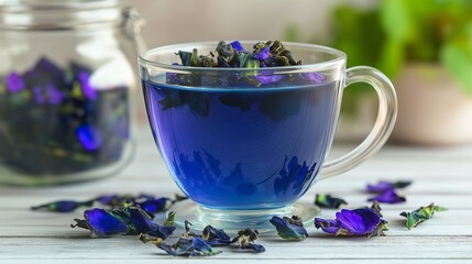 A cup of blue tea with flower petals in it. The cup is on a table with a jar of flowers next to it