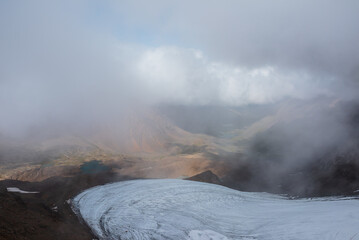 Misty aerial top view above round glacier to green rocky hilly valley with few turquoise mountain lakes under low clouds. Several most beautiful glacial lakes in low clouds in foggy changeable weather