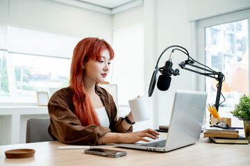 An attractive Asian female podcaster is working in her podcast studio, drinking coffee.