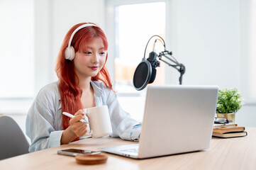 A beautiful Asian female podcaster is working in her home studio and enjoying a coffee.