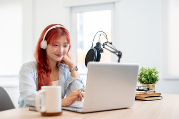 A happy Asian female podcaster is reading live comments on her online channel while broadcasting.
