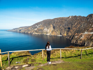 Fototapeta premium Young teenager girl explore beautiful nature scenes of Sliabh Liag viewing platform. Famous landmark in Ireland. Warm sunny day. Irish nature landscape. Travel and tourism industry.