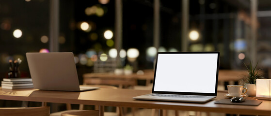 A laptop computer mockup on a wooden table in a contemporary coffee shop co-working space at night.