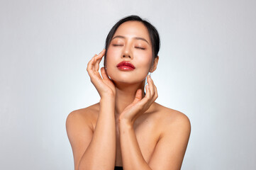 A beautiful Asian woman poses against a grey background, touching her face with her eyes closed.