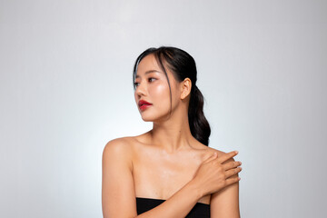 A gorgeous Asian woman touches her glowing, healthy tanned skin, posing against a grey background.