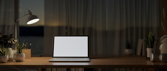 A laptop computer on a wooden table by the window in a house at night, illuminated by a table lamp.