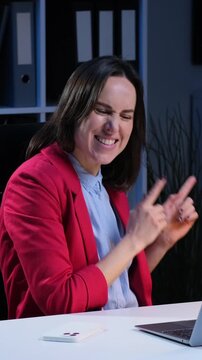 Smiley Caucasian female employee dancing and singing, sitting at desk in the evening office. Having fun during work break. Vertical video.