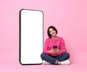 Beautiful young woman with smartphone sitting near big mobile phone with blank screen on pink...