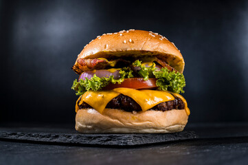 Delicious cheeseburger with lettuce, tomato, onion, bacon, and melted cheese, on a sesame seed bun, isolated on dark background. Mouth-watering and appetizing close-up.