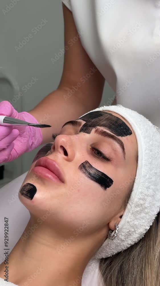 Wall mural cosmetologist applying black mask on pretty woman face wearing black gloves, gorgeous woman in spa h