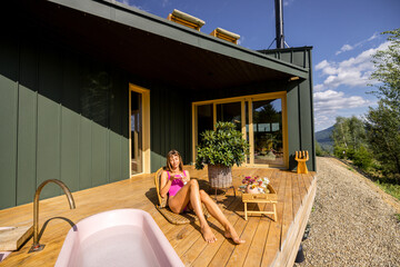 A woman enjoys a peaceful sunbath on a wooden deck, basking in the warm sunlight. Dressed in a vibrant pink swimsuit, she relaxes beside a pink bathtub, with a bottle of skincare nearby