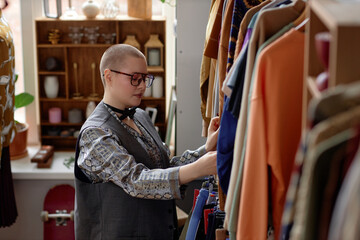 Side view of young female customer with buzz cut in vintage clothes thrifting at second hand shop...
