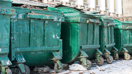 Row of large green metal dumpsters with peeling paint lined up outdoors near building. Ground around them is littered with debris and bins appear weathered and heavily used