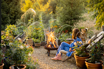 A woman enjoys a peaceful evening by the fire in a lush garden, surrounded by flowers and greenery. The setting sun casts a warm glow, enhancing the tranquil atmosphere of this outdoor retreat