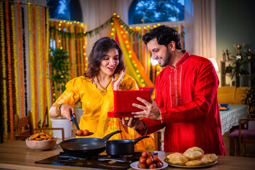 Indian young husband surprises wife with gift while cooking food on diwali festival eve in kitchen