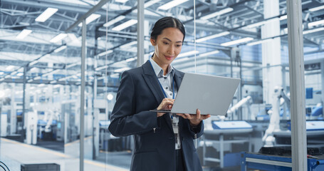 Fototapeta premium Asian Factory Manager Working on Laptop at Futuristic Industrial Manufacture with Autonomous Robotic Assembly Line. Specialist Monitoring Production, Planning Work for Workers