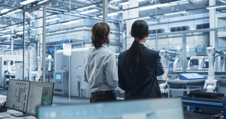 Fototapeta premium Young Caucasian Engineer and Asian Technician Having a Conversation and Using a Laptop Computer. Specialists Monitoring, Analyzing Conditions at a Modern Electronics Factory with Automated Conveyor